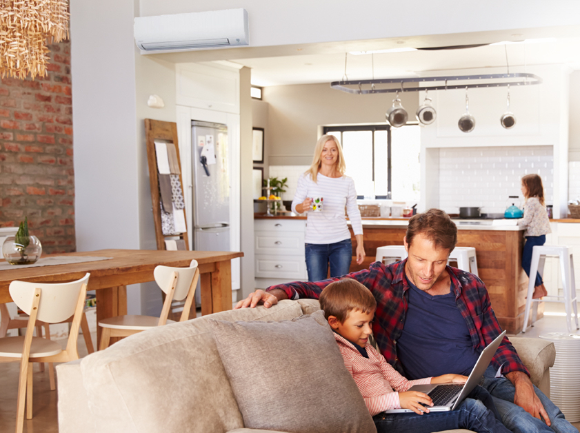 Father and son at home on a couch with mother and daughter in the background in the kitchen, with a heat pump on the wall.